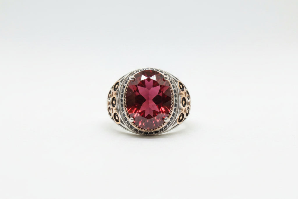 A silver-toned ring with a large oval red gemstone and small clear stones around it, displayed on a beige textured surface with price tags partially visible.