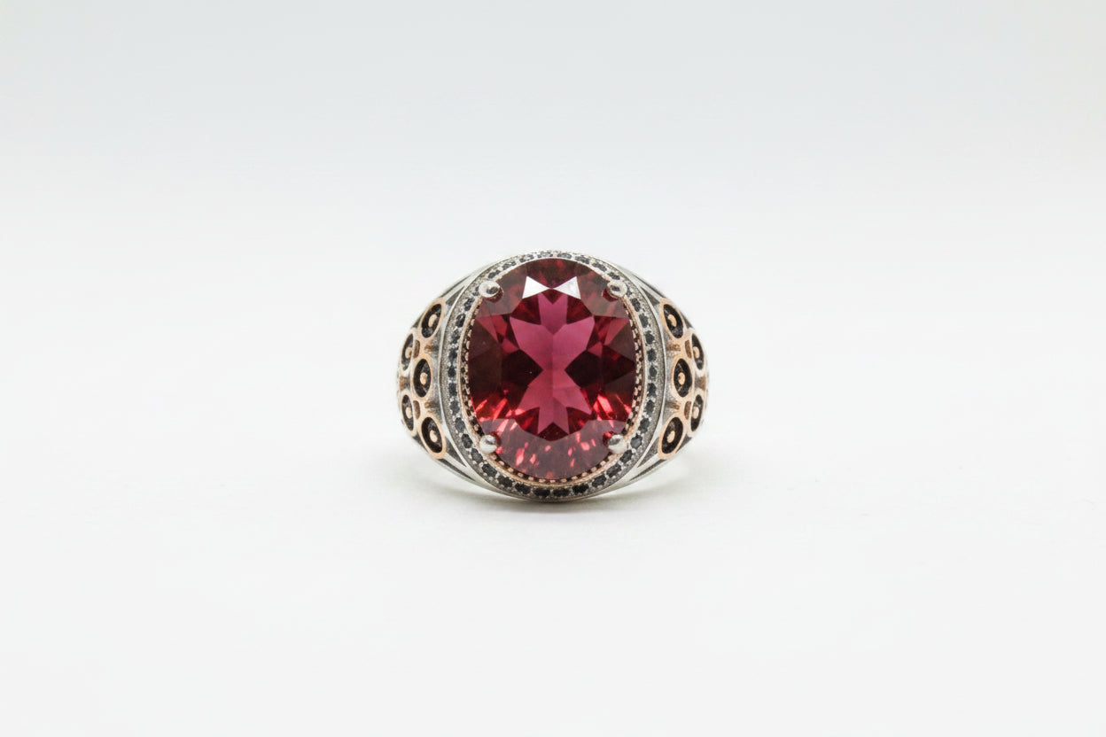 A silver-toned ring with a large oval red gemstone and small clear stones around it, displayed on a beige textured surface with price tags partially visible.