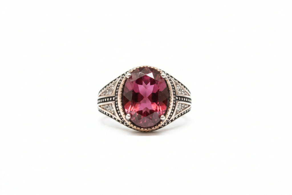 A silver-toned ring with a large oval pinkish-red gemstone, displayed on a beige textured surface next to a white price tag.